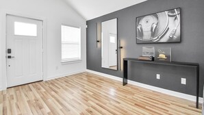 A hallway with a wooden floor, a white door, a window with blinds, a dark wall, and a console table with a framed picture and a hat. - Modern 3 Bedroom Close To DTWN/NRG/Med Center (Houston)