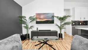 A modern living room with a glass coffee table, grey sofas, a black entertainment unit, and two potted plants. - Modern 3 Bedroom Close To DTWN/NRG/Med Center (Houston)