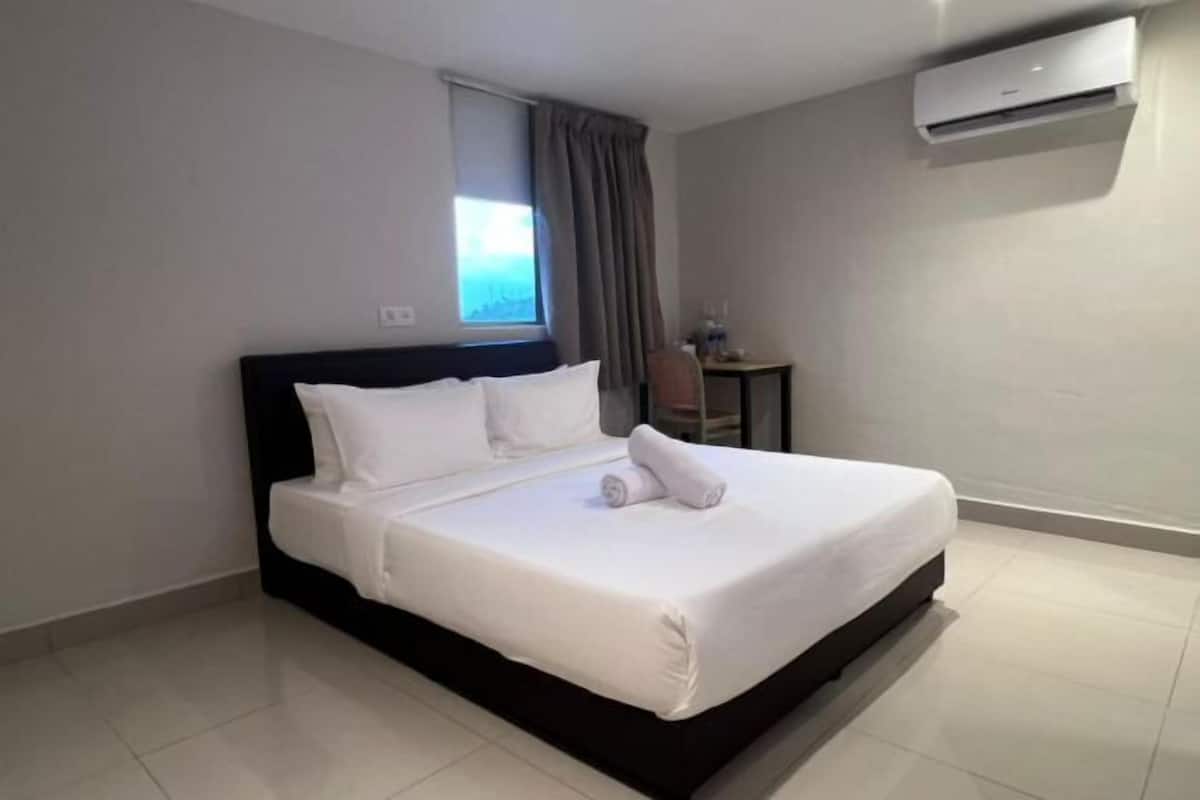 A hotel room with a bed, a window with curtains, a small table with a chair, and an air conditioning unit.