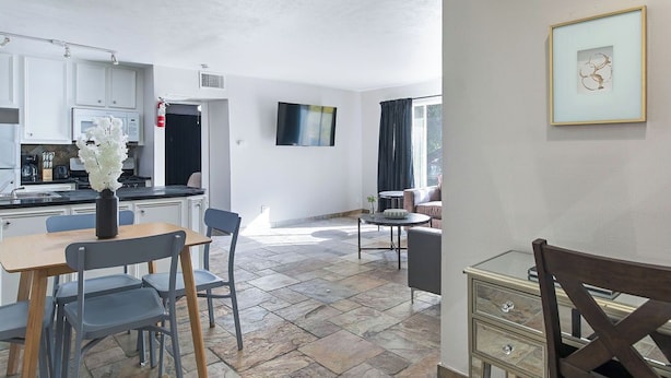 Una cucina moderna con mobili bianchi, un'area pranzo con tavolo e sedie, un televisore a parete e un'opera d'arte incorniciata.