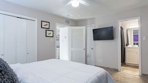 Una camera da letto con un letto, una ventola a soffitto, una TV e un bagno visibile attraverso una porta aperta.