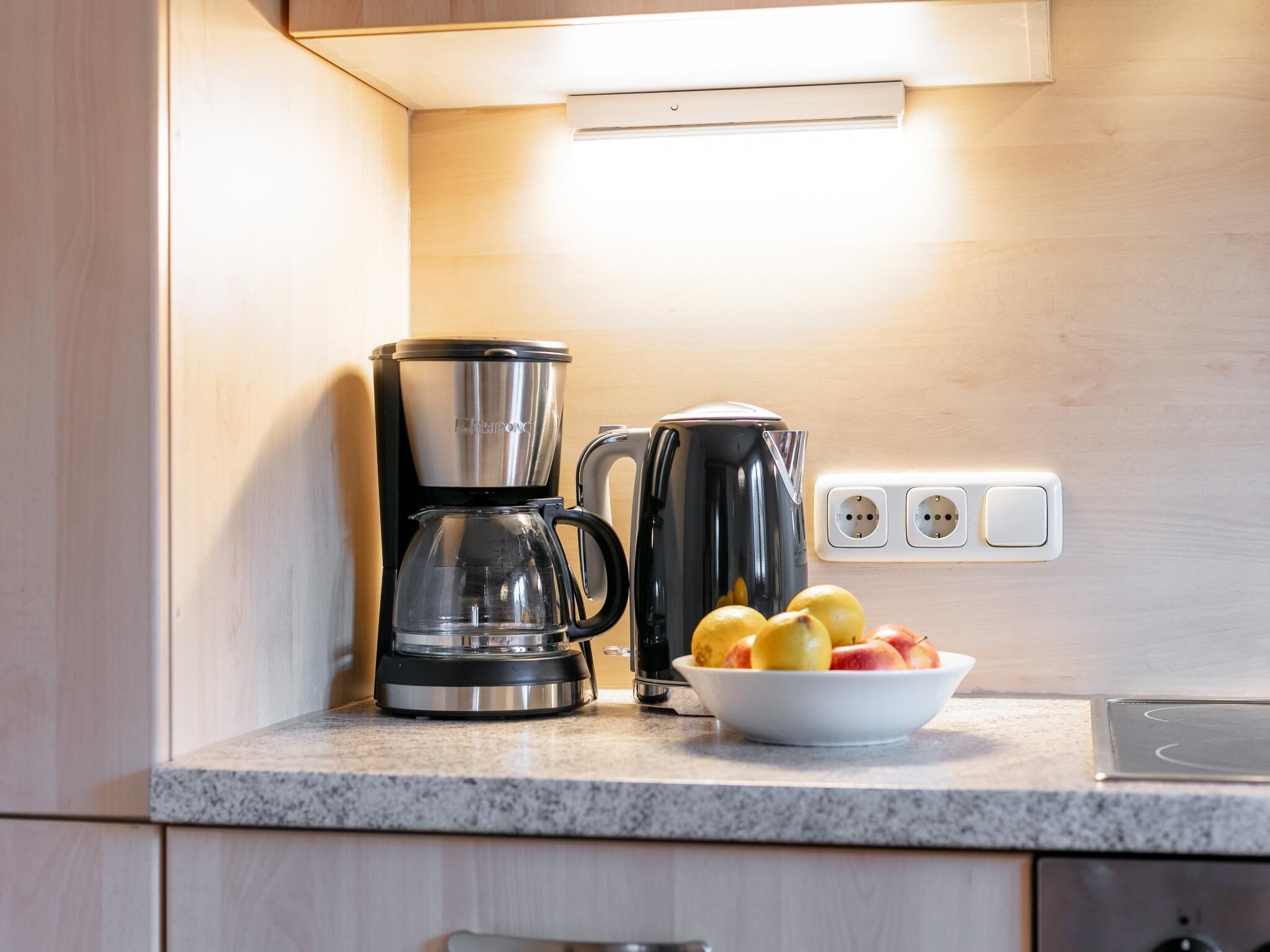 Una cocina con cafetera, hervidor y un bol de fruta en la encimera.