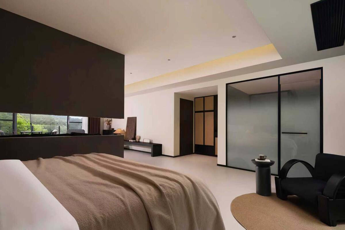 Een moderne hotelkamer met een groot bed, een zithoek met een tafel en stoelen, uitzicht op groen door het raam, en een schuifdeur naar een kast.