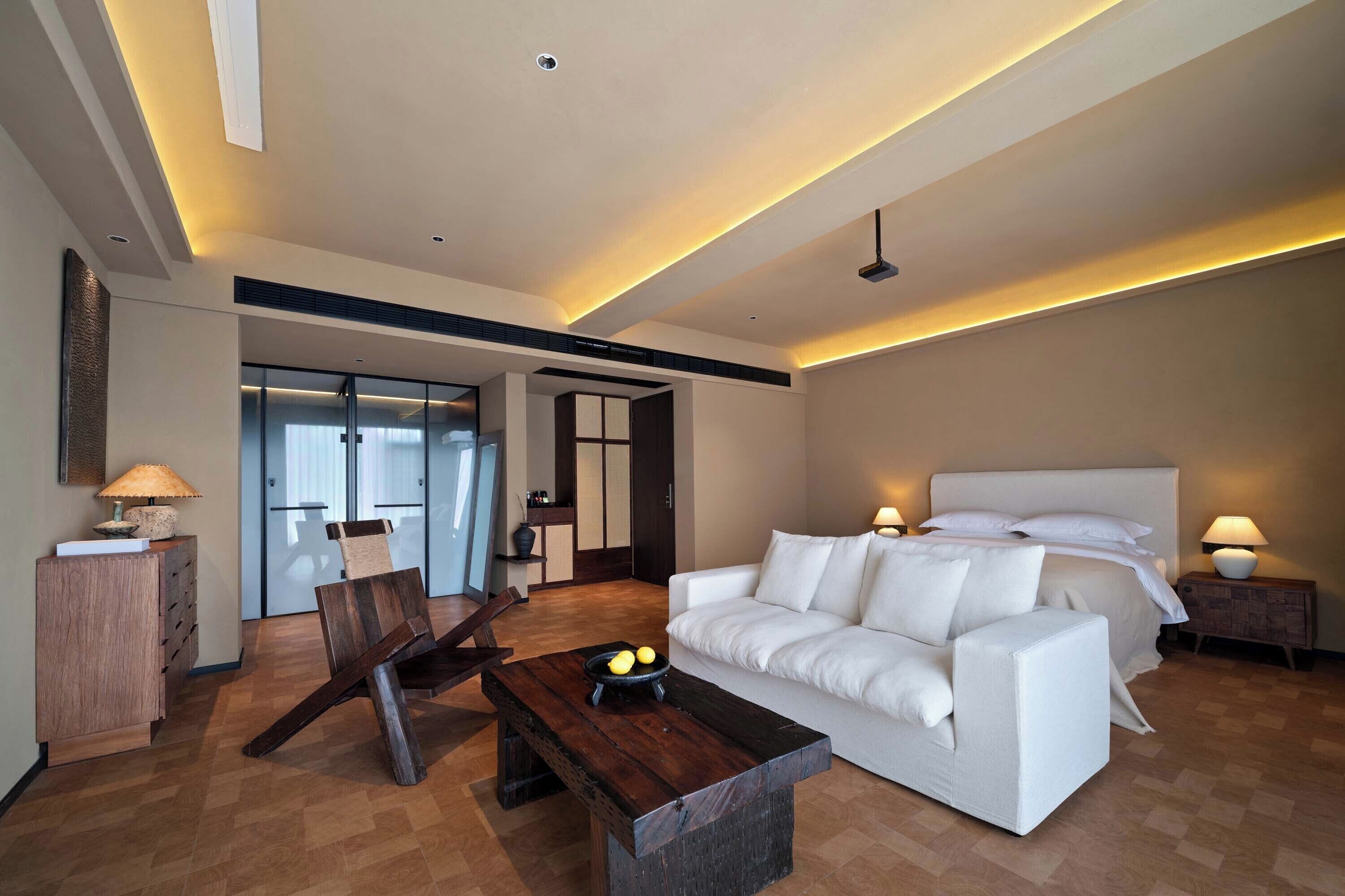 A modern hotel room with a large bed, a white sofa, a wooden coffee table, and a desk with a lamp.