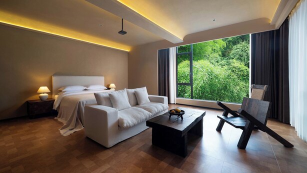 A modern hotel room with a large bed, a sofa, a coffee table, and a chair. There is a window with a view of greenery outside. - Fenghuang Border Town Mountain Homestay (Xiangxi)