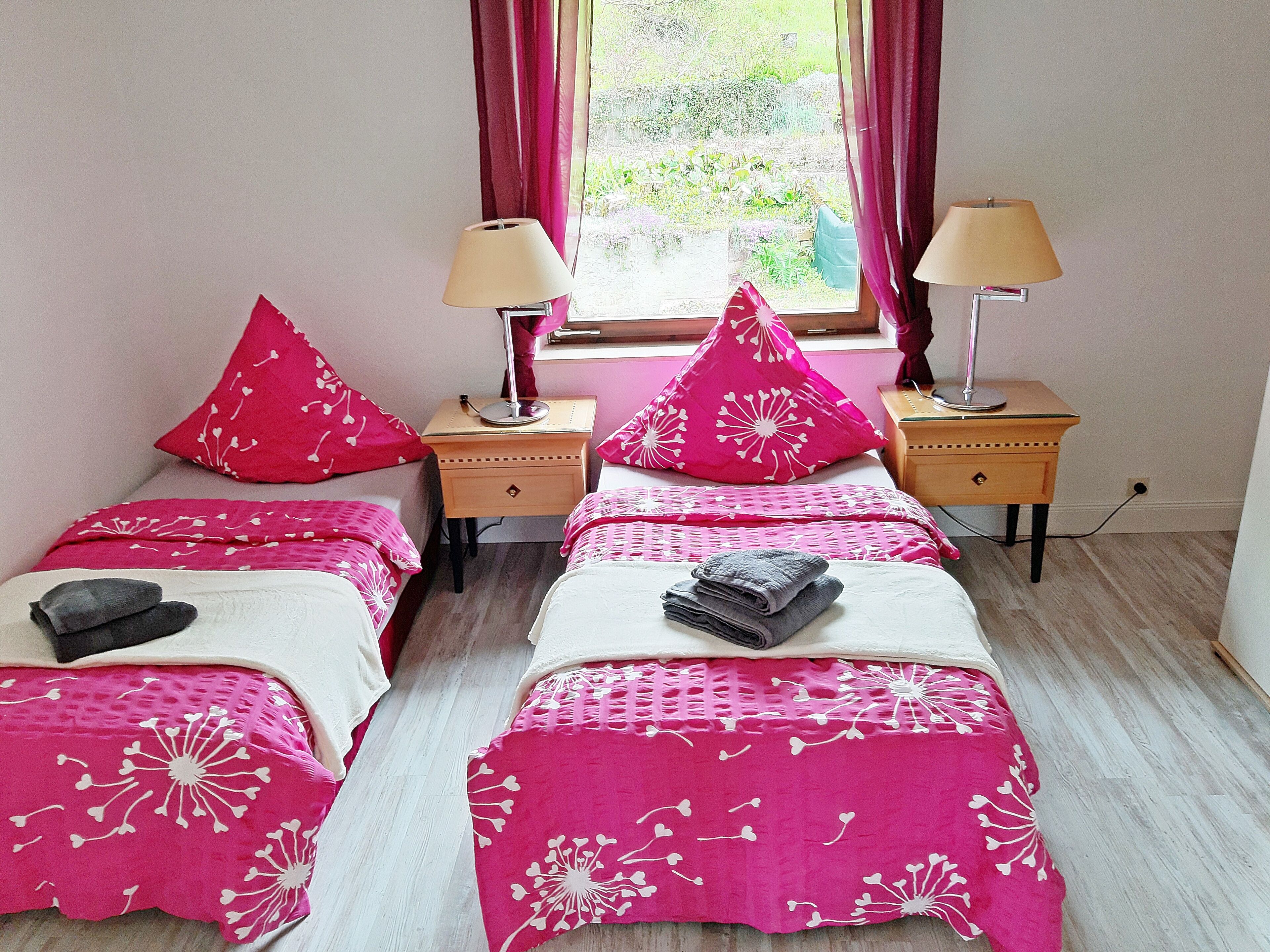 A room with two beds, each with a pink bedspread and matching pillows, a wooden nightstand with a lamp, and a window with curtains.