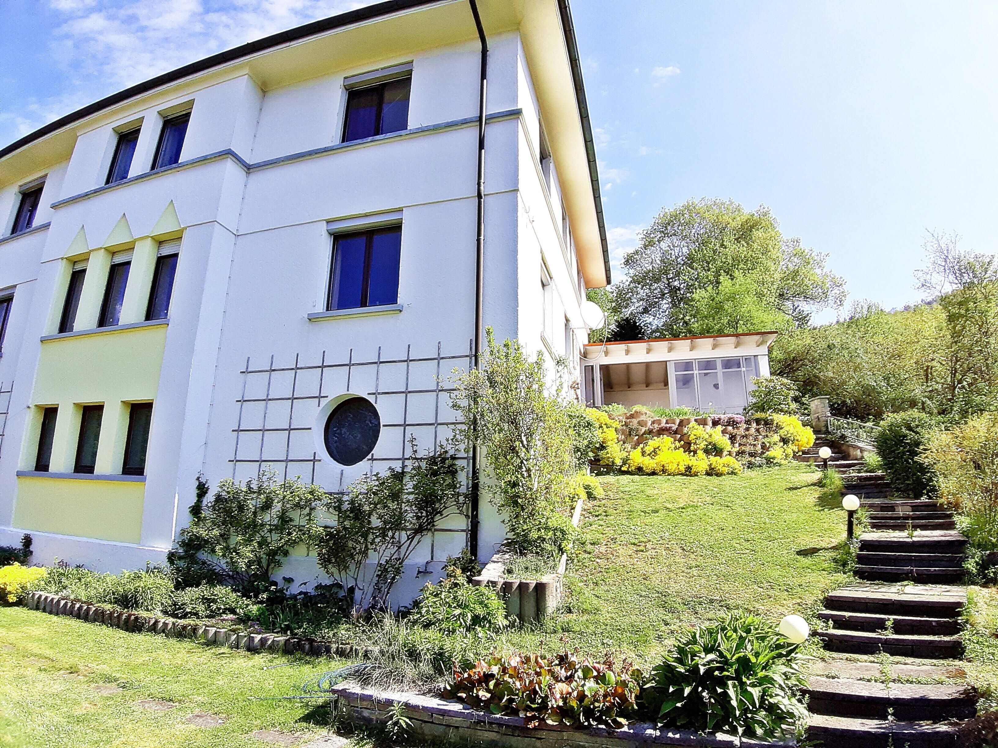 Un edificio de varios pisos con techo plano, ventanas amplias y un jardín cuidado con una escalera de piedra que lleva a una construcción pequeña.