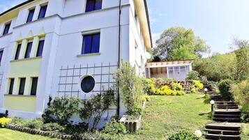 Un edificio de varios pisos con techo plano, ventanas amplias y un jardín cuidado con una escalera de piedra que lleva a una construcción pequeña.