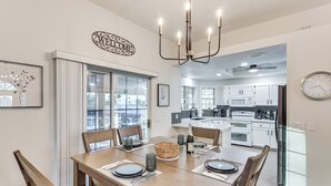 A modern dining area with a wooden table, chairs, and a chandelier. There is a kitchen visible in the background with white cabinets and a microwave. - Private Pool, Spa! Updated Home Near London Bridge (Lake Havasu City)