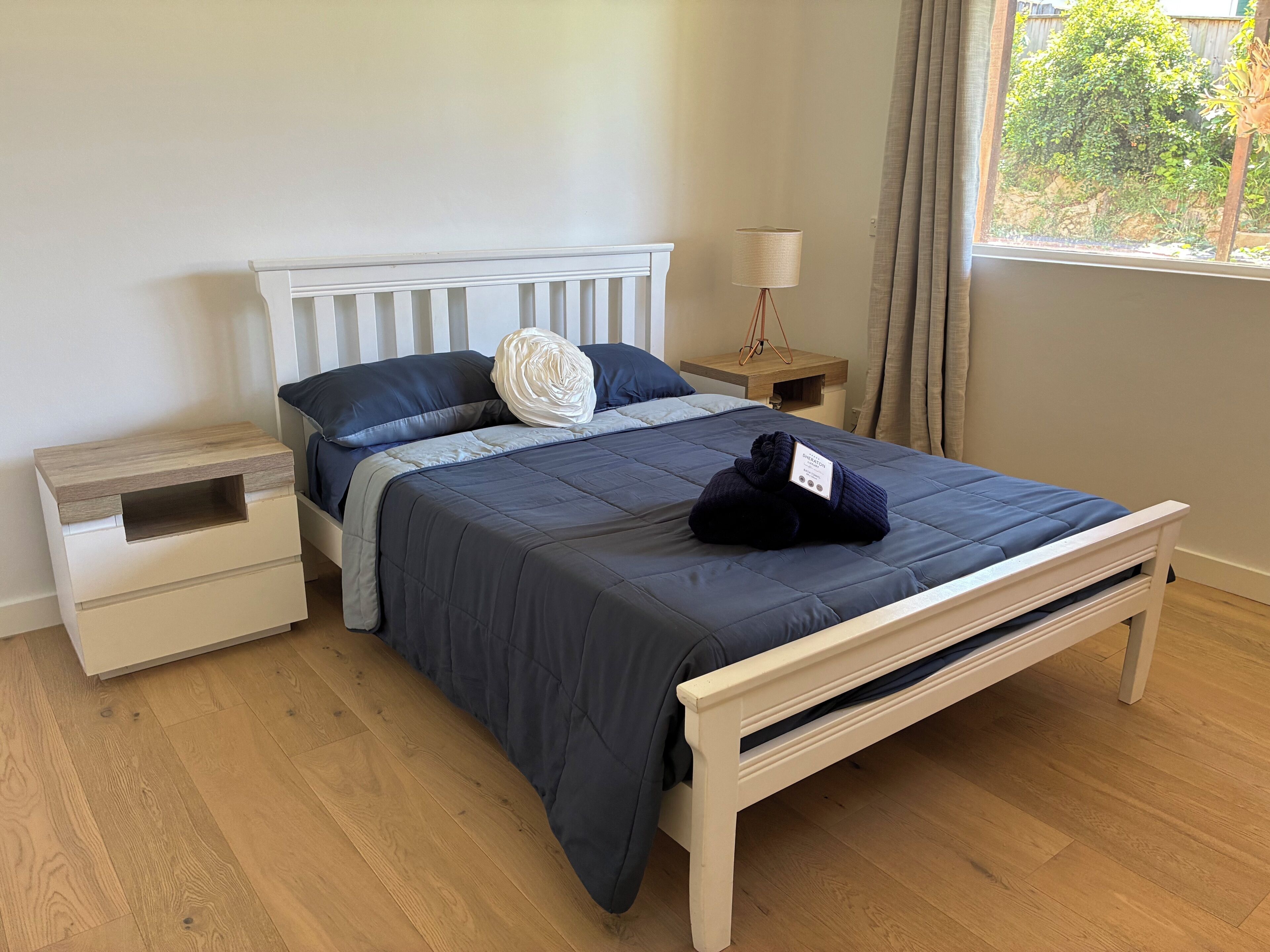 A neatly made bed with a dark blue bedspread, a white headboard, and a wooden nightstand with a drawer.