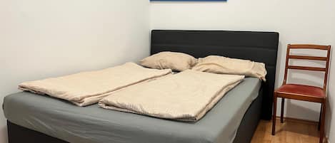 A neatly made bed with a grey mattress and beige bedding, a wooden chair, and a framed picture of sunflowers on the wall.
