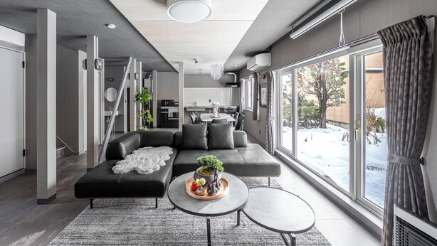 A modern living room with a black sectional sofa, a coffee table with a plant, and a view of a snowy outdoor area through large windows. - THE LAVENRIDGE (Nakafurano)