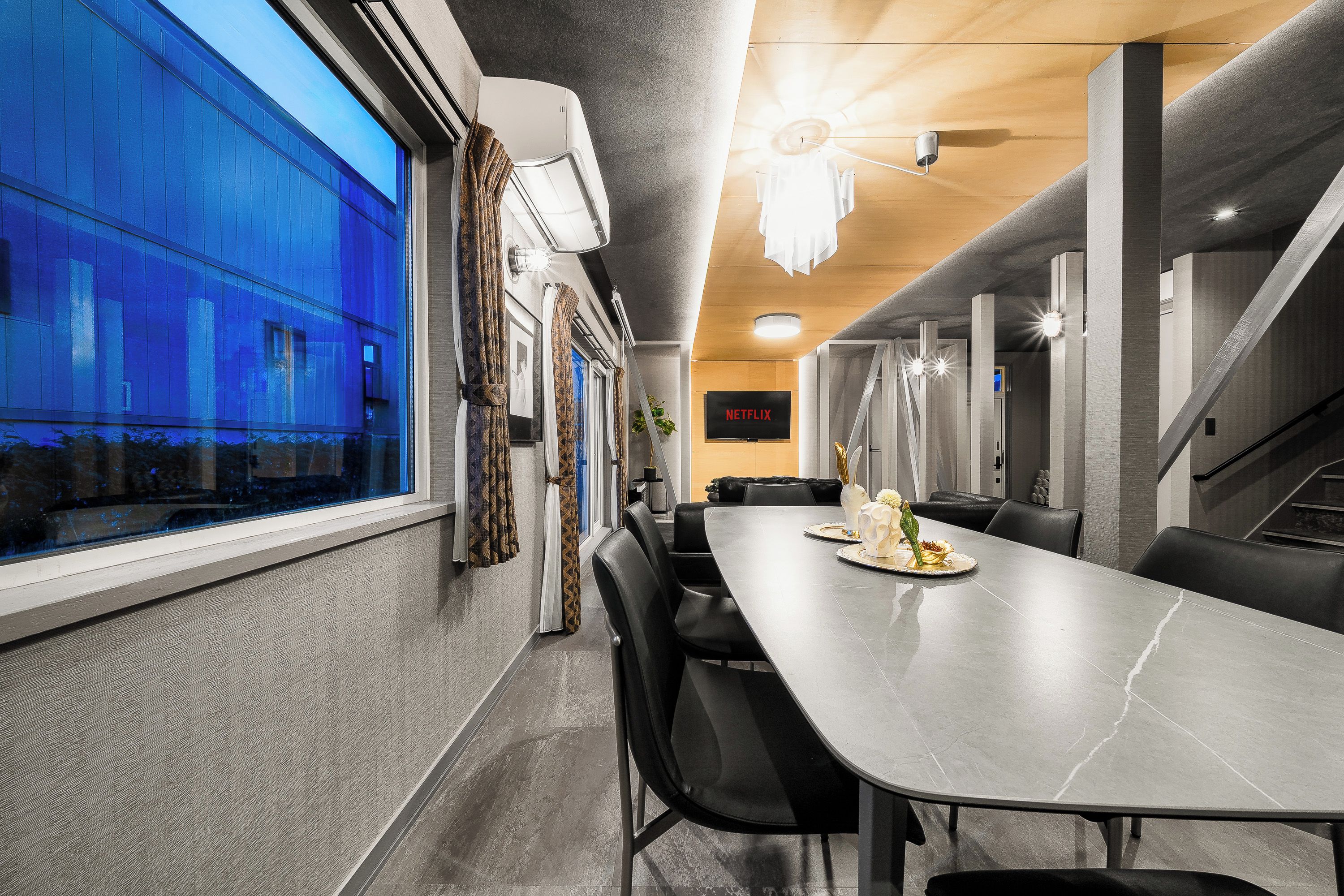 A modern dining area with a long table, chairs, and a view of a building through large windows.