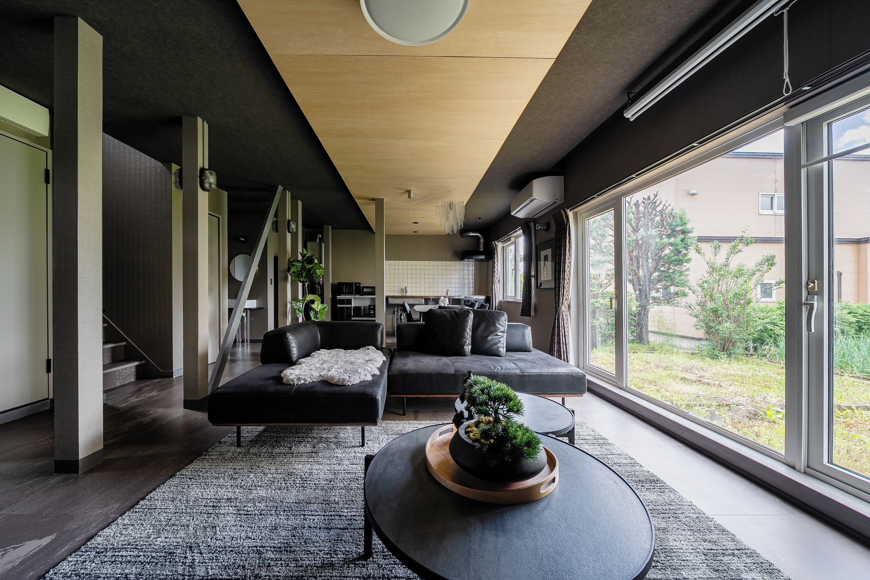 A modern living room with a sofa, a coffee table with a plant, and a view of the outdoors through large windows.