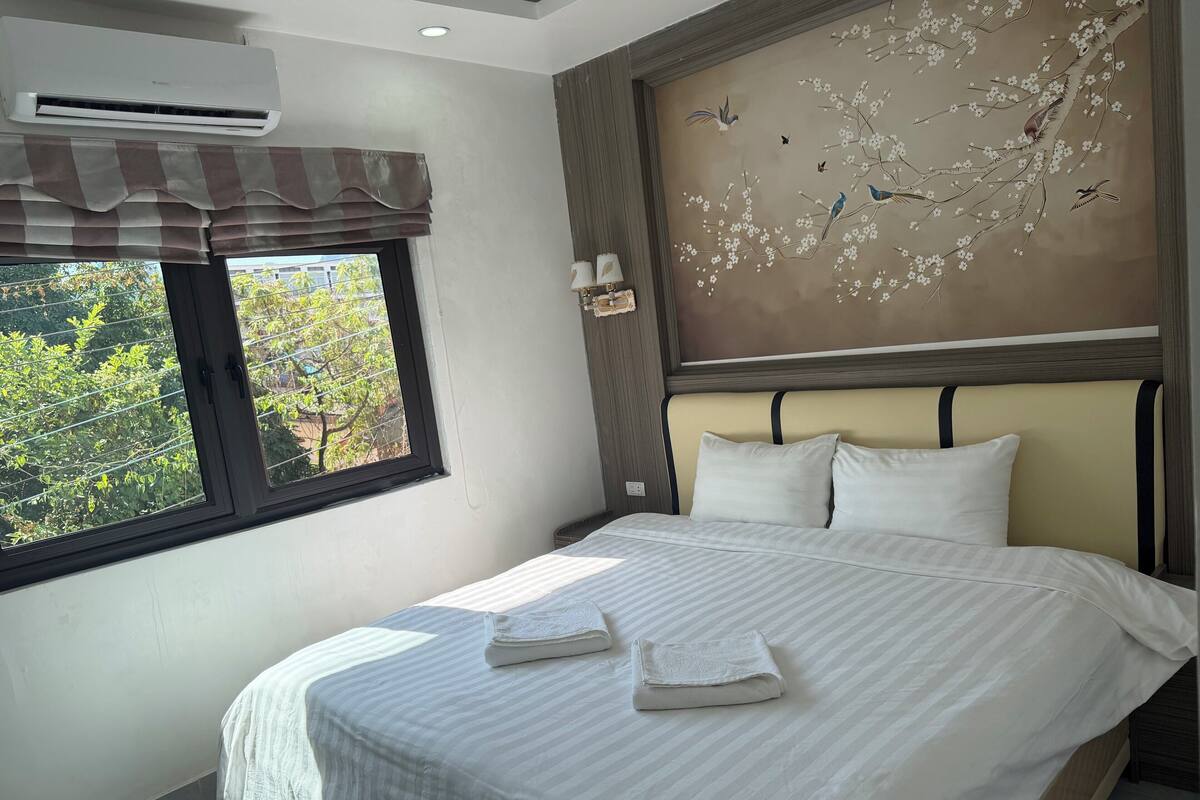 A neatly made bed with white linens and two folded towels, a window with a view of greenery, and a wall with a floral mural.