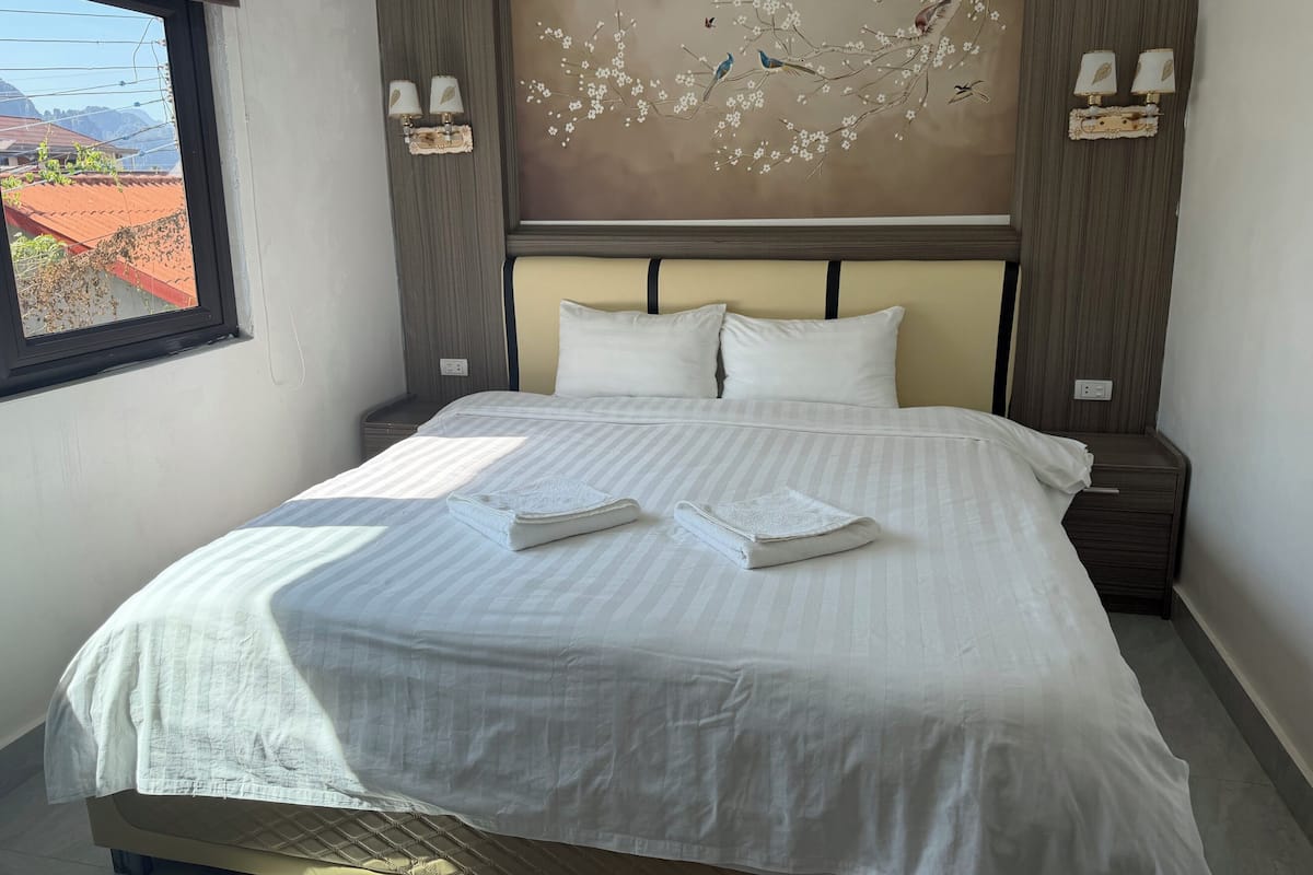 A neatly made bed with white linens, two pillows, and two folded towels. There are wall-mounted lamps on either side of the bed.