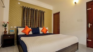 A neatly made bed with a white and navy blue bedspread, two orange pillows, and a wooden headboard. There is a nightstand with a vase of flowers and two water bottles beside the bed. A wooden door and a wall-mounted light fixture are visible in the room. - Affordable garden plus view stay in misty Munnar (Chithirapuram)