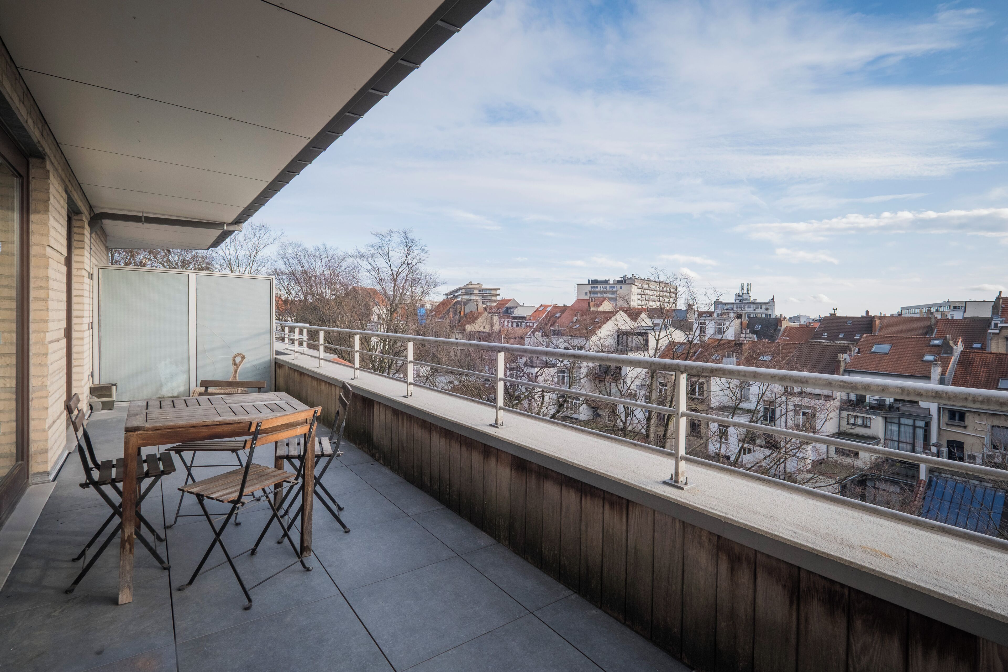 Un balcon avec une table et des chaises, donnant sur un paysage urbain avec des immeubles et un ciel dégagé.