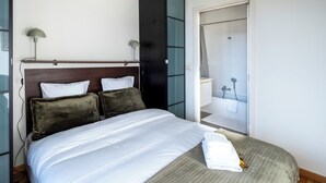 A neatly made bed with white linens and two dark pillows, a bedside lamp, and a view of a bathroom with a shower and toilet. - Brussels EU Center residence (Brussels)