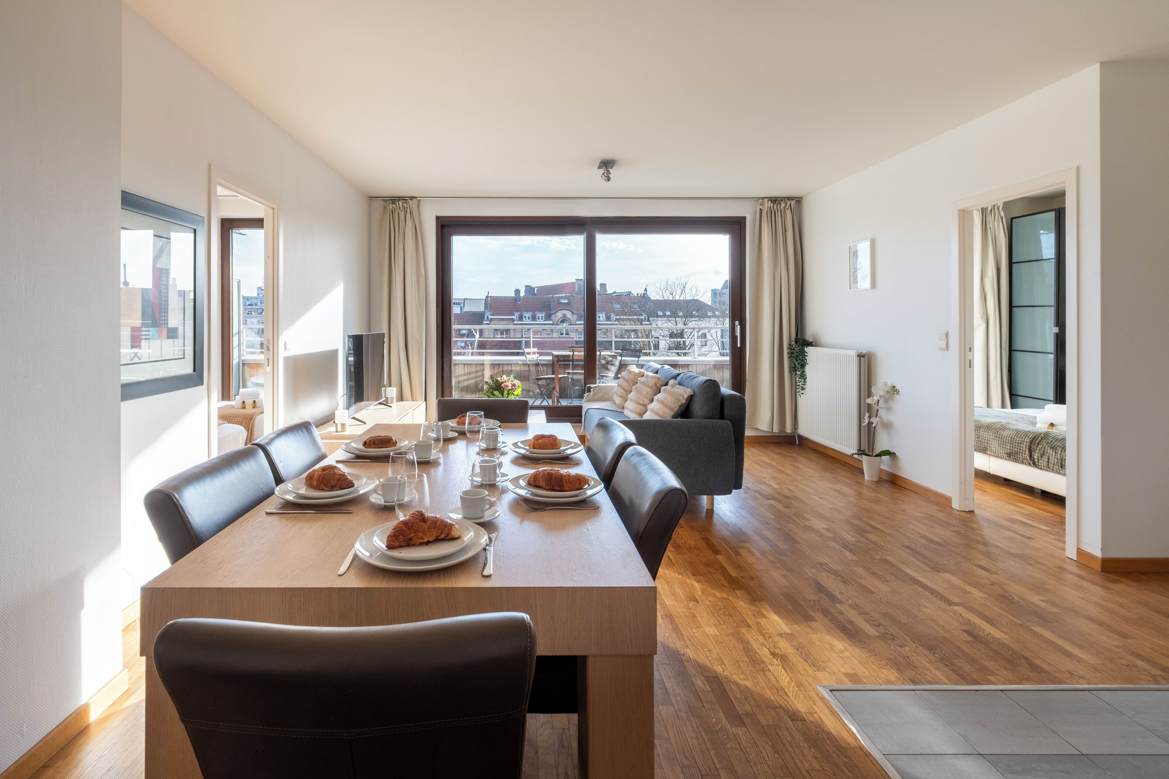 Un comedor con una mesa preparada para cuatro personas, un sofá y vistas de la ciudad a través de amplios ventanales.