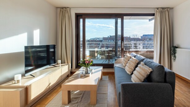 A modern living room with a flat-screen TV, a sofa, a coffee table, and a large window offering a city view. - Brussels EU Center residence (Brussels)