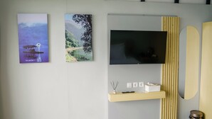 A wall with two framed landscape paintings, a flat-screen TV, a wooden shelf with a remote and a small vase, and a mirror.