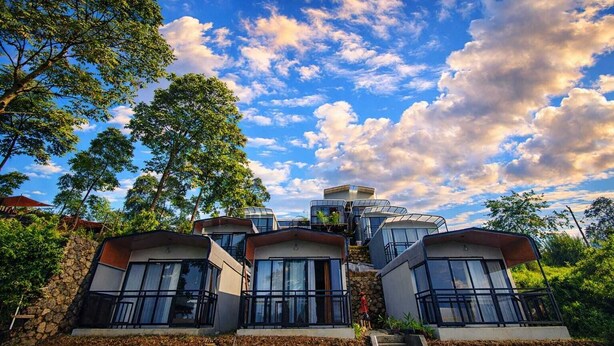 A modern house with multiple levels and large windows, surrounded by trees and a clear sky.