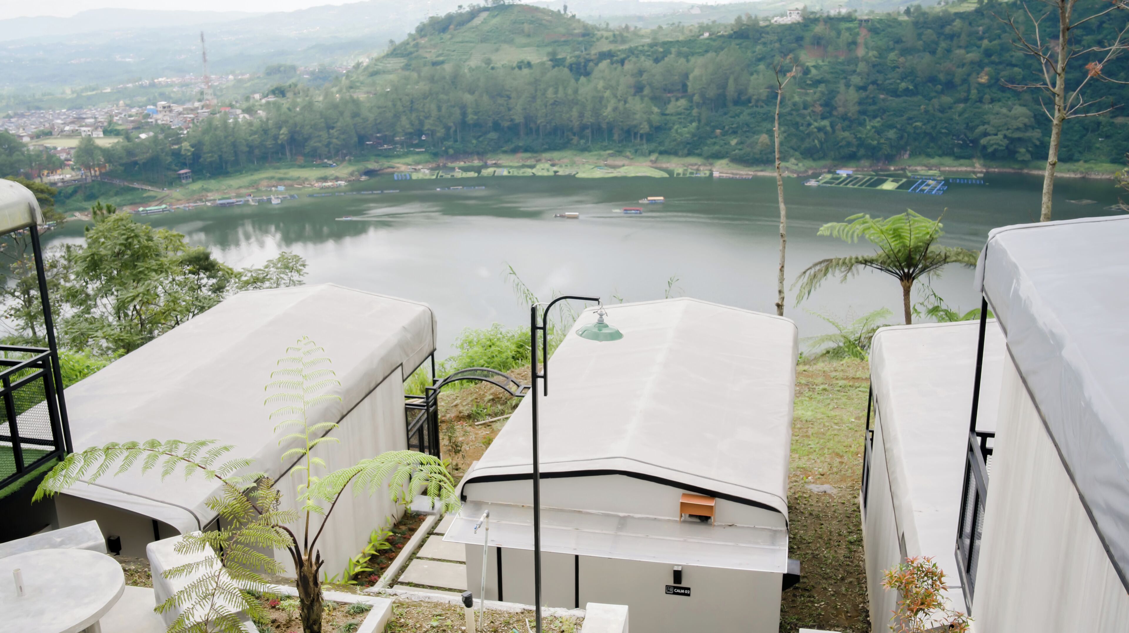 Vista de un lago con barcos, rodeado de vegetación y colinas. En primer plano, se observan estructuras cubiertas con una mesa y sillas.