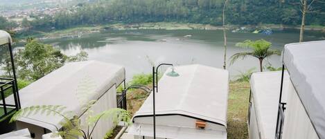 Vista de un lago con barcos, rodeado de vegetación y colinas. En primer plano, se observan estructuras cubiertas con una mesa y sillas.