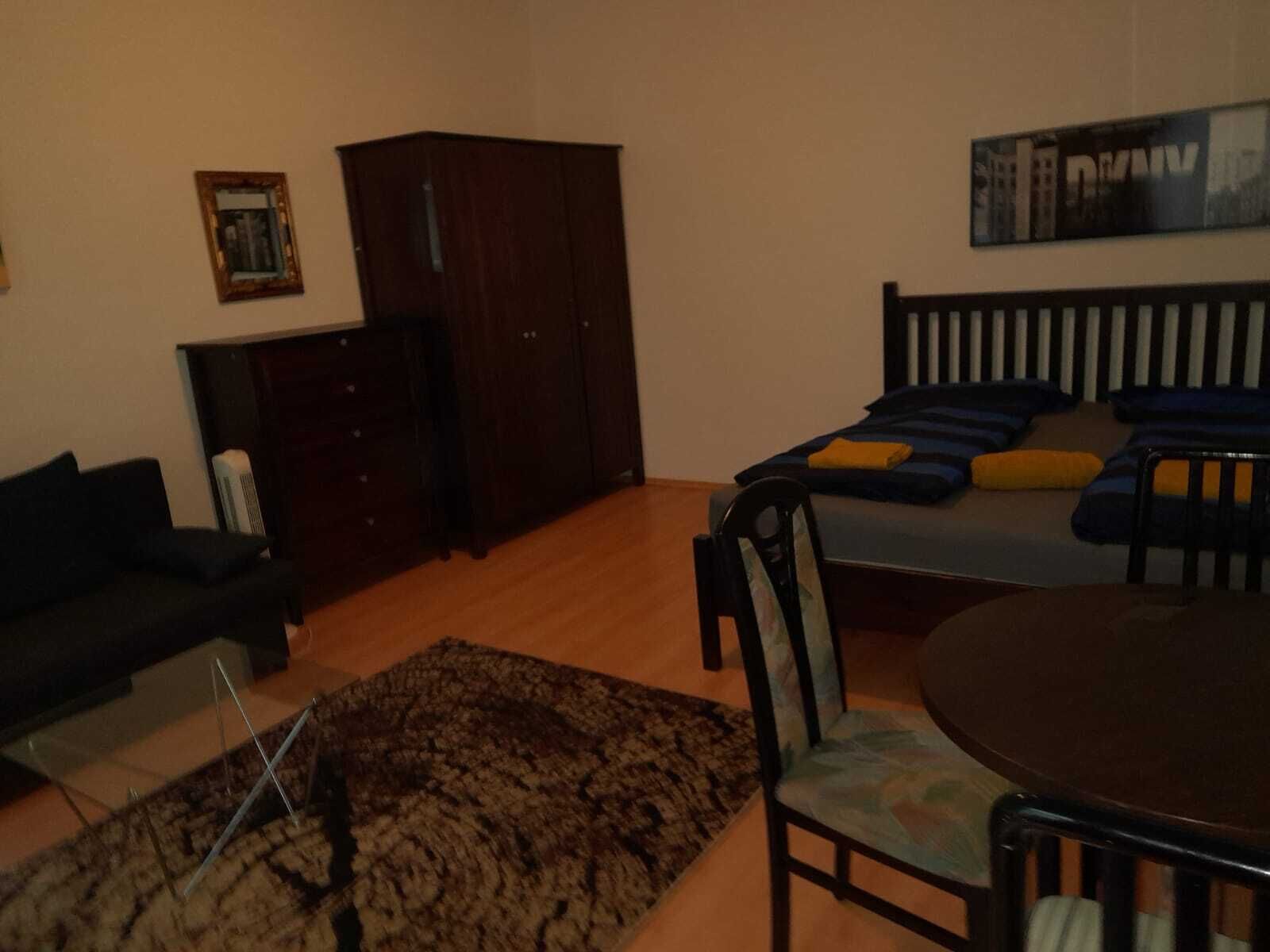 Quarto com guarda-roupa de madeira escura, cama com almofadas azuis e laranja, mesa redonda pequena e sofá de cor escura.