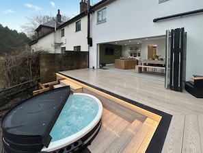 Un espace moderne aménagé à l’extérieur avec un jacuzzi, une terrasse en bois et un espace de repas clos par des parois de verre, le tout surmonté d’un bâtiment blanc en arrière-plan.