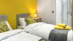 Quarto de hotel com uma cama, uma mesa de cabeceira com abajur e um box de chuveiro de vidro.