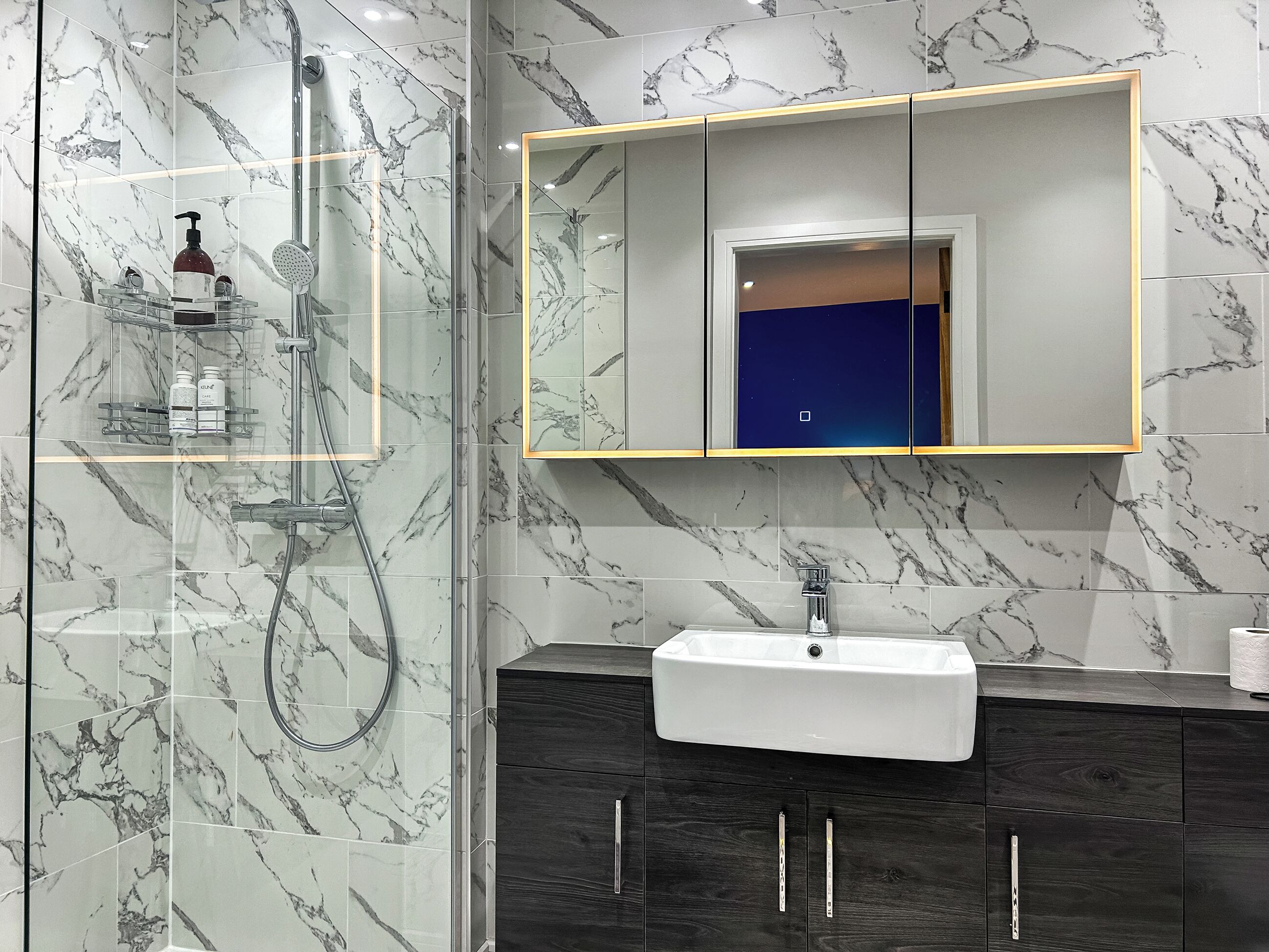 A bathroom with a glass-enclosed shower, a white sink, dark wooden cabinets, and a marble-tiled wall.