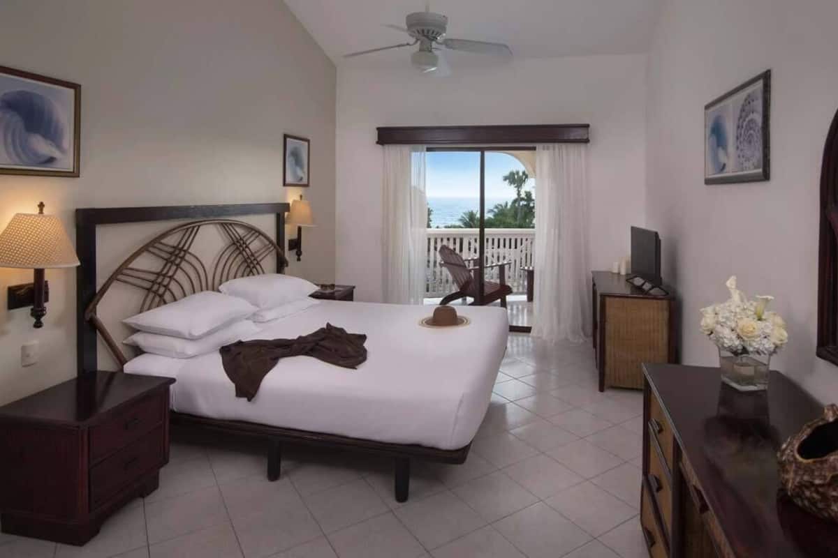 Een hotelkamer met een bed, nachtkastje, dressoir, televisie en een balkon met uitzicht op zee.