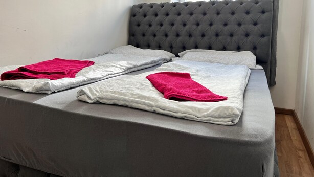 A neatly made bed with grey bedding and two red towels, a grey tufted headboard, and a window with curtains. - Comfortable 3BR Apartment in Vienna (Vienna)