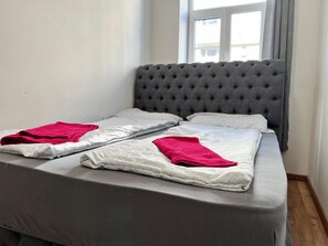 Ein ordentlich bezogenes Bett mit grauem Bettzeug und zwei roten Handtüchern, ein grauer, mit Wirbeln versehener Kopfteil sowie ein Fenster mit Vorhängen.