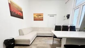 A modern, minimalist room with a beige sectional sofa, a white desk, and two black chairs. There are two paintings on the walls, a door, and a window with blinds. - Comfortable 3BR Apartment in Vienna (Vienna)