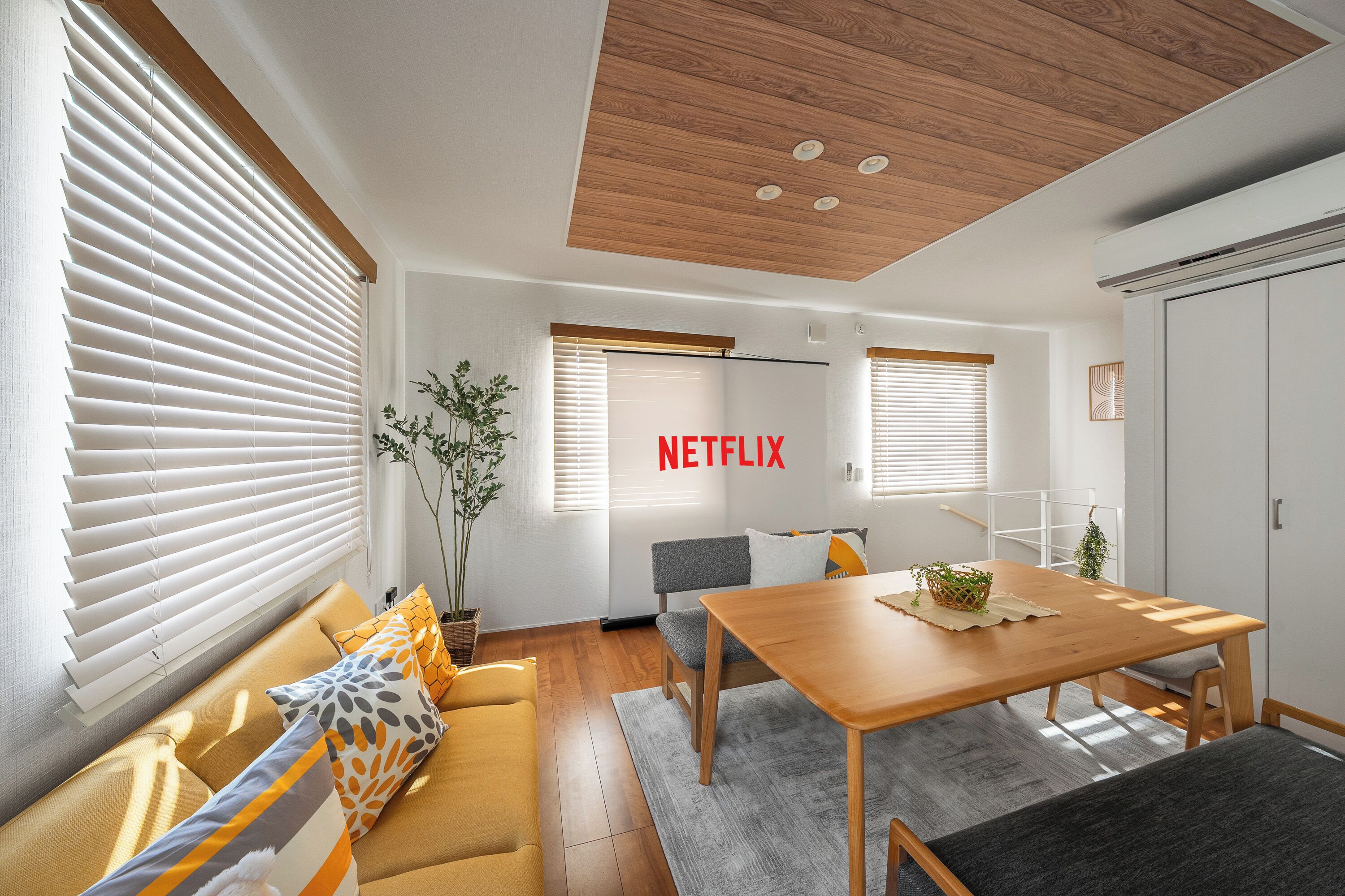 A modern living room with a sofa, a wooden dining table, and a TV screen displaying the word "NETFLIX".