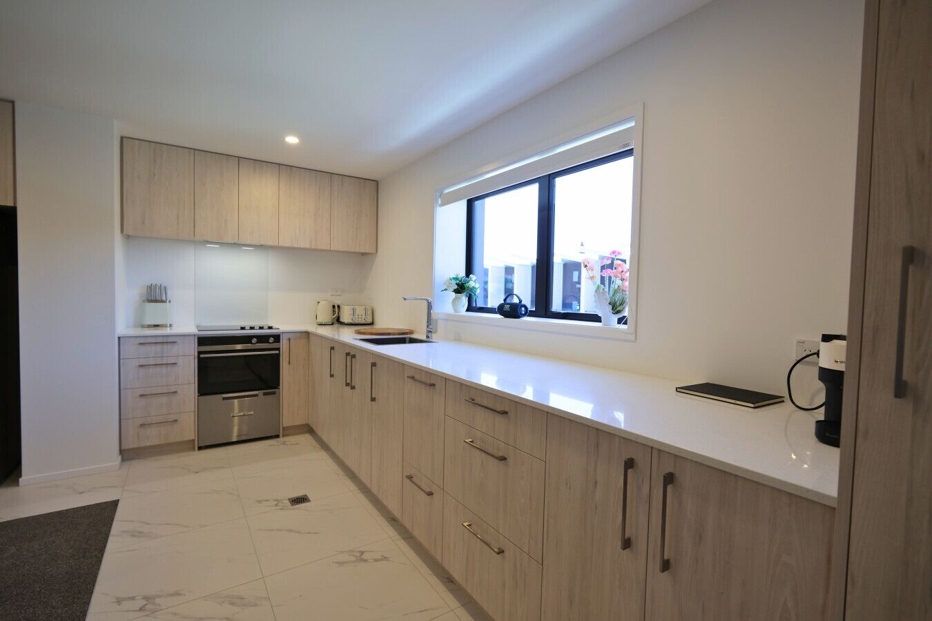 Een moderne keuken met lichte houten kastjes, een oven van roestvrij staal en een raam met uitzicht op gebouwen.