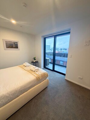A hotel room with a large bed, a small table with a plant, a framed picture on the wall, and a view of the city through a large window. - Brand-New 1BR Apartment near Te Kaha Stadium (Christchurch)