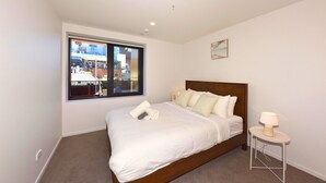 A bedroom with a large window, a bed with white bedding, a bedside table with a lamp, and a small painting on the wall. - Newly Built 1BR Apartment near Te Kaha Stadium (Christchurch)