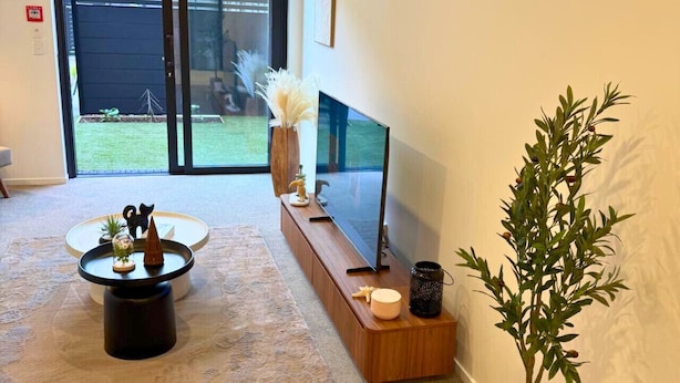 A modern living room with a large flat-screen TV, a wooden coffee table, a potted plant, and a sliding glass door leading to a backyard. - Central 1BR Apartment Steps From Te Kaha Stadium (Christchurch)