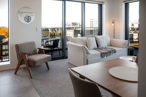Un salon moderne comprenant un canapé, un fauteuil, une table à manger et une grande fenêtre donnant sur des immeubles.