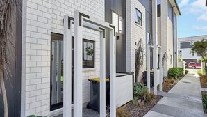 A modern multi-story residential building with white brick and grey siding, a covered outdoor seating area, and a paved walkway. - Stylish 2-Bedroom Home with Back Yard (Christchurch)