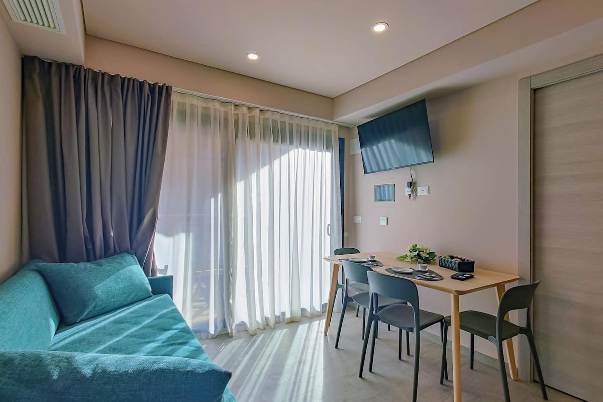 A modern room with a sofa, a dining table with chairs, a TV mounted on the wall, and a window with curtains.