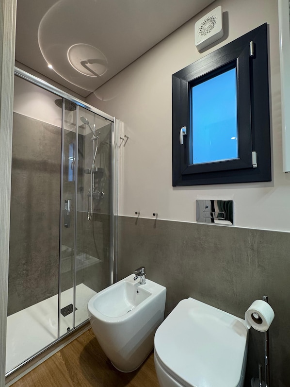Uma casa de banho moderna com chuveiro em cabine de vidro, um bidé, uma sanita e um espelho com janela azulada.
