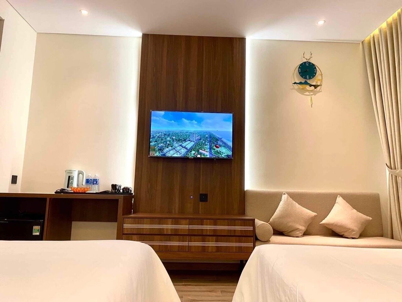 Hotellihuone, jossa on suuri, seinälle asennettu litteänäyttöinen televisio, sänky, jossa on kaksi tyynyä, yöpöytä, jossa on vedenkeitin ja kuppeja, sekä seinäkello.
