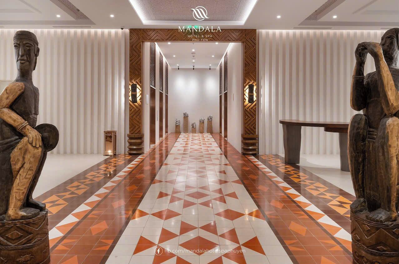 A hallway with a geometric patterned floor, wooden statues on either side, and a central entrance with the hotel name 'MANDALA HOTEL & SPA PHU YEN'.