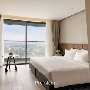 Una habitación de hotel con una cama grande, una mesita con un florero y vistas a un paisaje urbano a través de un ventanal amplio.