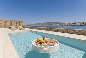 Ein Pool mit einem schwimmenden Frühstücksbrettchen, Steinmauern und Blick auf das Meer.